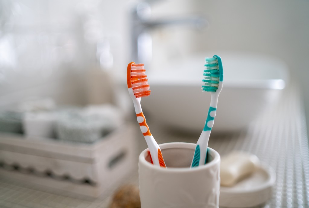 Two colorful toothbrushes stand in a ceramic glass against the background of the washbasin and other accessories in the bathroom.